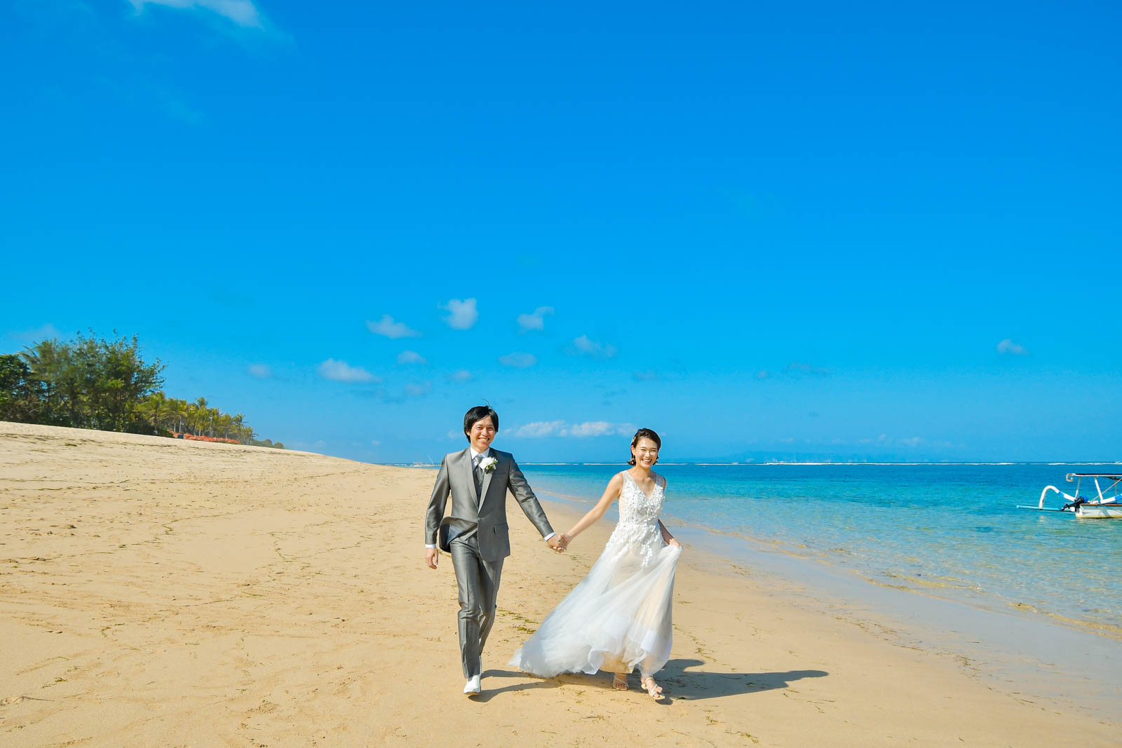 青い海を背景に、砂浜を手をつないで歩く新郎新婦のビーチフォト。軽やかなドレスが風に揺れるシーン